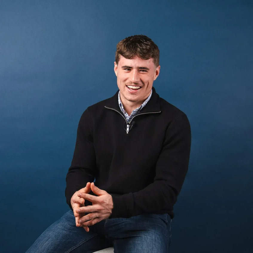 Man wearing a black sweater over a patterned shirt against a blue background