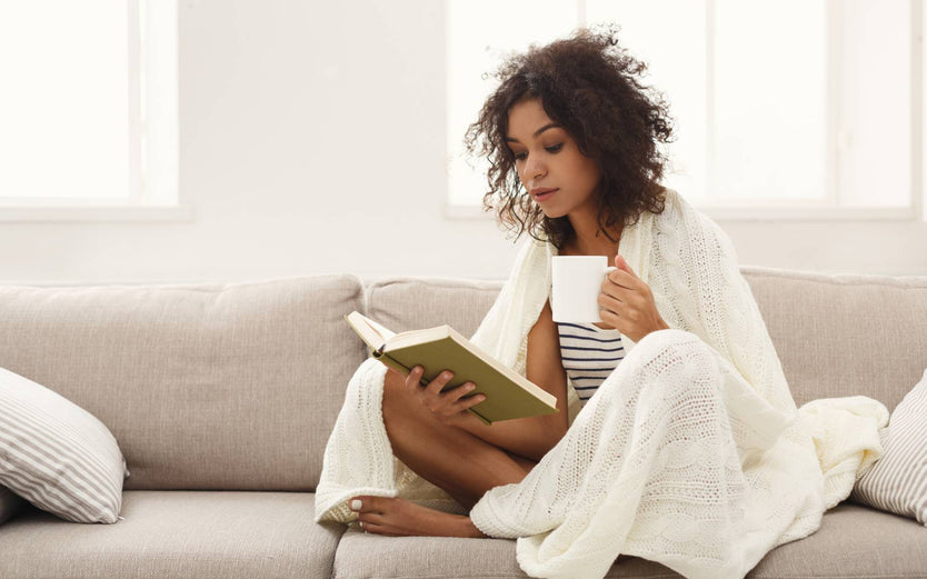 Woman sitting under a blanket on a couch reading a book and drinking a cup of tea