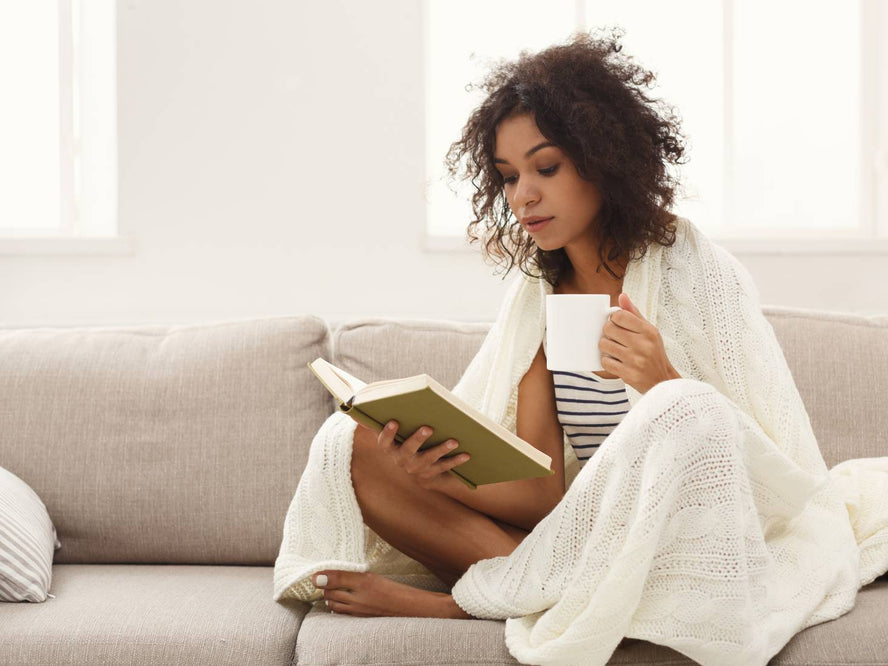 Woman sitting under a blanket on a couch reading a book and drinking a cup of tea