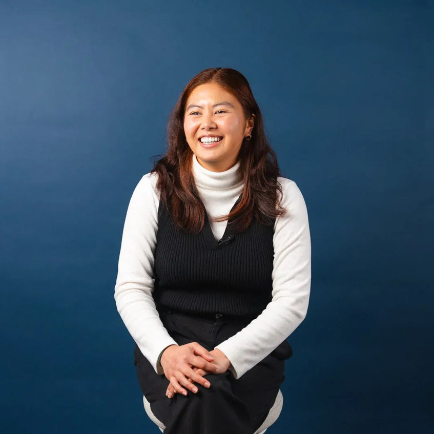 Woman sitting against a blue background wearing a white turtleneck and black vest.