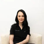 Woman wearing a black shirt with a logo, sitting on a white couch against a white wall.