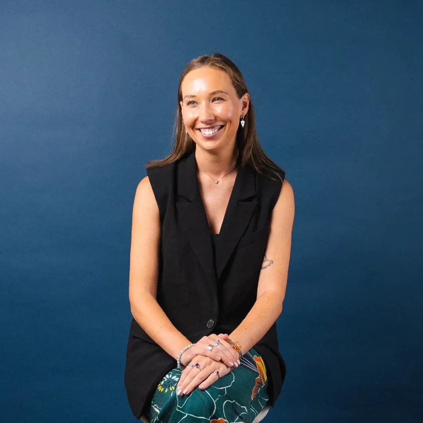 Woman sitting against a blue background wearing a black sleeveless blazer and colorful pants.