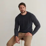 Man wearing a dark blue sweater and beige pants sitting on a stool against a plain background