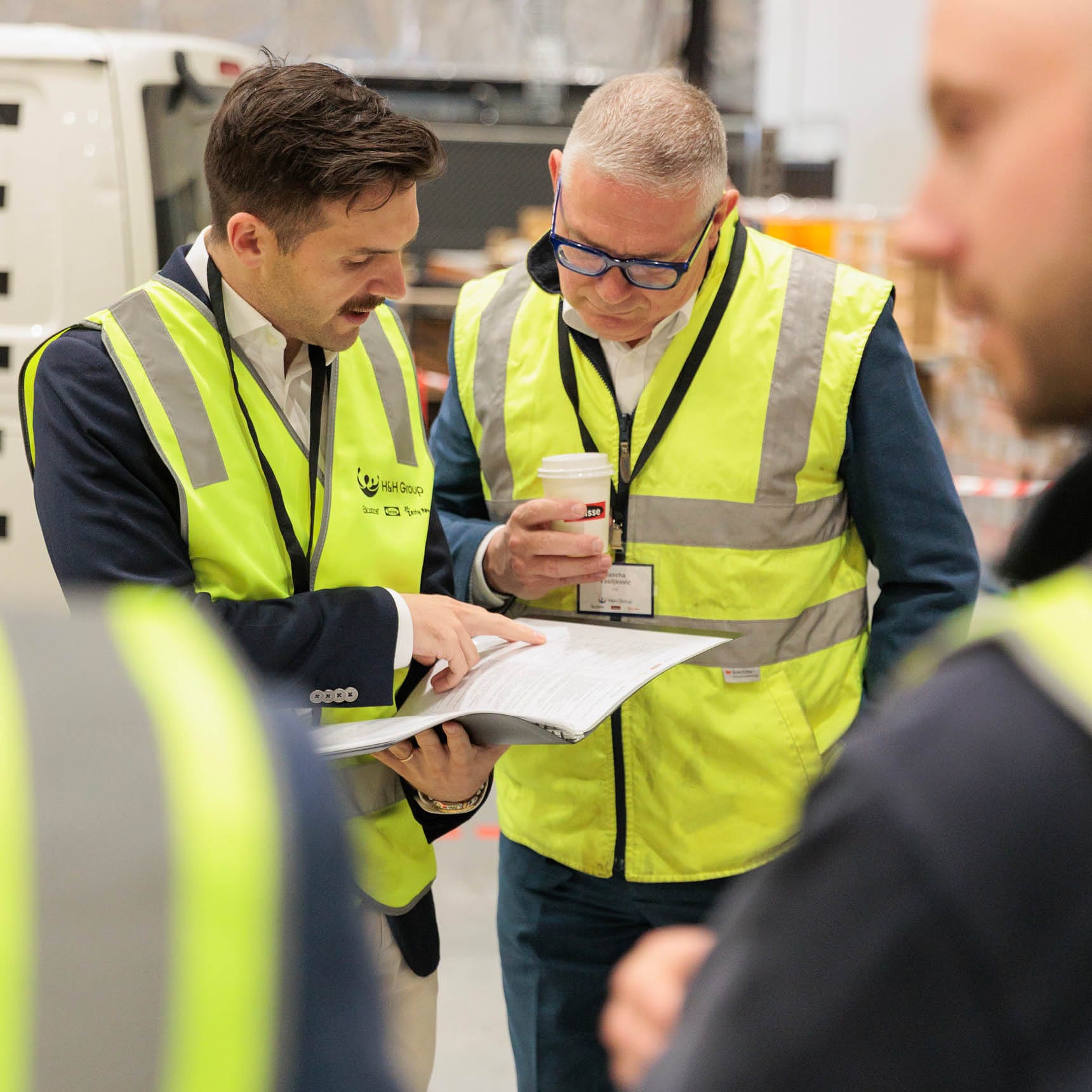 Two men in high-visibility vests reviewing documents in a warehouse setting.