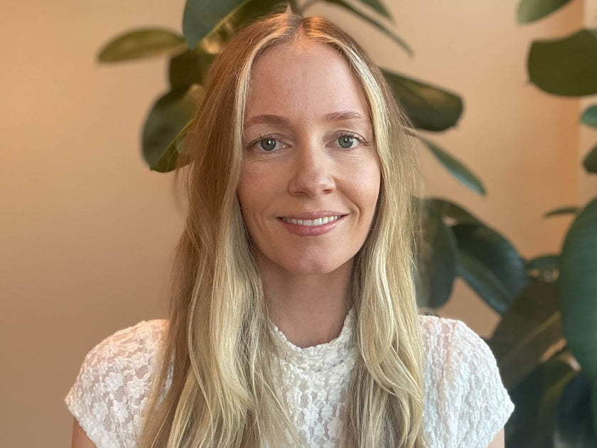 Woman with blonde hair wearing a white lace top in front of green plants.