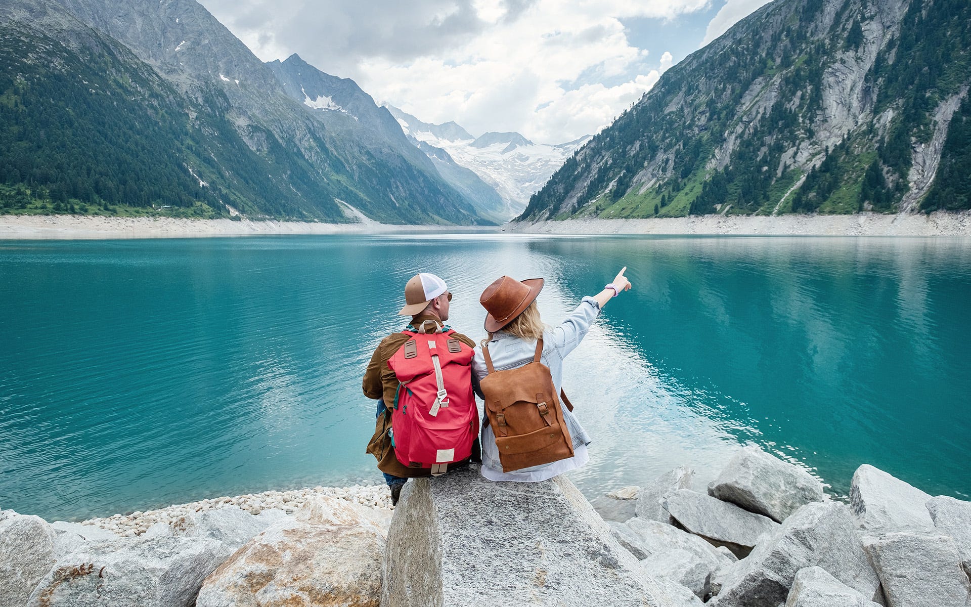 Two people sitting in front of a lake surrounded by mountains.